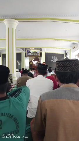 A group of Muslim men are gathered in a mosque for prayer. They are all facing towards the front of the mosque, where an imam is likely leading the prayer. The mosque appears to be a modern building with white walls and a tiled floor. There are columns supporting the ceiling and a large screen at the front displaying an image. The men are dressed in traditional Islamic attire, including caps and robes. Some of the men are seated on the floor, while others are standing. The atmosphere appears to be one of reverence and devotion.