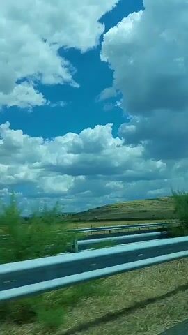 The video shows a view from a moving vehicle, likely a car, traveling along a highway. The perspective is from the passenger side, looking out the window. In the foreground, there's a silver metal guardrail. Beyond the guardrail is a grassy area with some low-lying green shrubs or bushes that appear to be swaying in the wind. In the mid-ground, there's a vast expanse of green fields, possibly agricultural land, stretching towards rolling hills in the distance. The sky is a vibrant blue, dotted with large, fluffy white clouds, suggesting a sunny day with some cloud cover. The overall scene depicts a rural landscape during daytime.