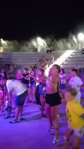 A group of people are dancing on a stage at night. The stage is lit with purple lights. The people are wearing casual clothing. Some of the people are children and some are adults. There is an audience sitting in bleachers in the background. There is also some mist or smoke in the air.