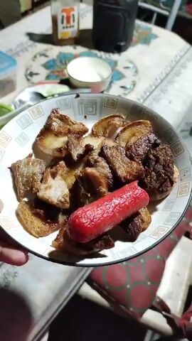 The video shows a close-up view of a plate of food, primarily consisting of several pieces of fried pork belly and a hot dog. The food is served on a white plate with a decorative pattern. In the background, there is another plate of sliced cucumbers and red onions, with a fork and spoon on it. There are also other items visible on a table, including a white bowl, a bottle, and some green vegetables. The lighting suggests an indoor setting, possibly a mealtime.