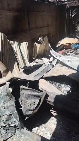 The video shows the interior of a building that has been damaged, possibly by fire. The ground is covered with debris, including corrugated metal sheets that appear to be burnt. There is a concrete wall on one side, and a staircase or framework visible through an opening at the end of the space. A white barrel, some boxes, and other scattered items can be seen in the background. A person says "Esta es la casa de mi hermana," which means "This is my sister's house" in Spanish. The scene suggests a recent destructive event, leaving the building in ruins.