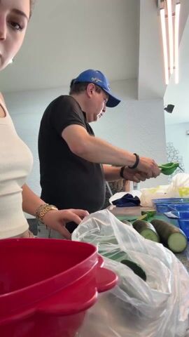 A woman and a man are standing at a kitchen counter preparing food. The woman is on the left, wearing a white tank top, and is cutting a cucumber. The man is on the right, wearing a black t-shirt and a blue baseball cap, and appears to be scooping something with a green utensil. There are several cucumbers on the counter near them, still in a plastic bag. In front of the woman is a large red bowl. Behind the man, there are blue containers. The room is well-lit with modern lighting fixtures. There are other people in the background, but they are out of focus. The wall behind the man has a textured, wavy design.