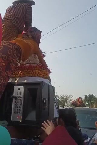 The video shows an outdoor event, possibly a parade or festival. In the foreground, a person in traditional clothing is leaning over a large, decorated structure, possibly a float. They are handing items, which appear to be chilies, down to someone below. Another person in traditional clothing is visible behind them, wearing a conical hat and also engaged in the activity. A large speaker is mounted on the structure, and a group of people are gathered below, some with their hands raised. Indonesian flags are also visible. The sky is clear and bright.