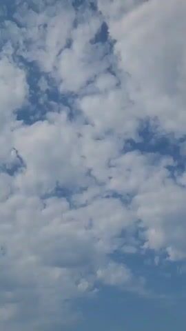The video shows a slightly tilted perspective looking up at a bright blue sky filled with scattered white cumulus clouds. The camera appears to be moving slowly upwards, revealing more of the sky as it progresses. There are no people or objects visible in the frame, only the natural phenomenon of clouds against the sky. The lighting suggests a daytime scene, likely sunny with a moderate amount of cloud cover.
