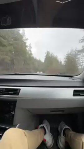 The video shows a car's dashboard and a driver's legs and feet as they drive down a tree-lined road on a rainy day. The car has a silver dashboard and black floor mats, and the driver is wearing tan pants and white sneakers with red accents. Other vehicles are visible on the road ahead. The windshield wipers are active. There appears to be camping gear strapped to the roof of the car.