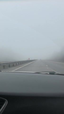 The video depicts a car driving on a highway engulfed in heavy fog. The visibility is significantly reduced, with the road ahead barely discernible. Guardrails are visible on the left side of the road, and there are a few other vehicles ahead, including a truck. The windshield wipers are on, suggesting moisture is present on the glass. The interior of the car is visible, and music is playing in the background.