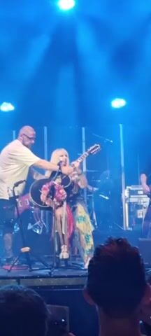 A man in a white t-shirt and black shorts stands to the left of a woman seated on a stool on a stage. The woman has long blonde hair and is wearing a colorful dress. She is playing a 12-string acoustic guitar, which is decorated with flowers. A microphone is positioned in front of her. Another man wearing a dark shirt is visible behind the woman, holding a red electric guitar. The stage is lit with blue lights, and a backdrop of vertical panels is behind the performers. The audience is visible in the foreground.