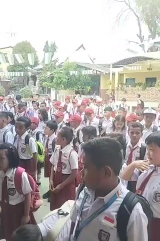 The video shows a large group of elementary school children in uniform, standing in an outdoor schoolyard. The children are wearing white shirts, maroon skirts or shorts, and red ties. Many have backpacks. The children appear to be listening to someone, possibly a teacher, as indicated by the verbal interaction. The background includes school buildings, trees, and some decorative banners. The overall impression is of a school assembly or a gathering before or after class.