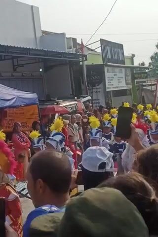 A marching band parade is taking place outdoors on a street. The band members are wearing blue and white uniforms with yellow plumes on their hats. Some are holding instruments, while others are marching. A crowd of spectators is gathered to watch the parade. The background shows buildings and tents, indicating a commercial or community event. The weather appears to be sunny.