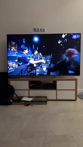 The video opens with a wide shot of a living room, focusing on a large television displaying an esports event. A man in a blue shirt is visible on the left side of the screen, holding a microphone and speaking. The game on the TV transitions to a title screen with "Intel Extreme Masters Cologne Major 2026" displayed prominently against a blue, crystalline background. The game then shifts to a first-person shooter gameplay scene, showing virtual combat with health bars and player names on display. A small inset window shows a player's face, identified as "RASPEEK". The TV is situated on a wooden and white media console with several shelves and cabinets. On the floor in front of the console, there are some items including a black backpack, a tape measure, and some cardboard packaging. The room has light-colored tiled flooring and neutral-colored walls.