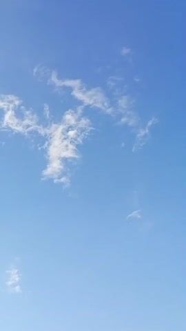 A vertical video captures a clear blue sky with scattered wispy white clouds. The clouds are small and delicate, with some appearing more textured and defined than others. The overall impression is of a serene, open sky. There are no people, objects, or discernible actions in the frame.