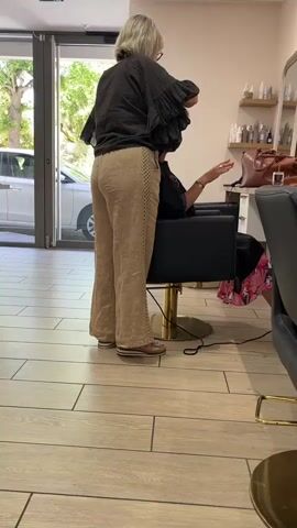 A blonde woman in beige pants and a dark top is standing behind a person sitting in a black chair. The blonde woman is holding a black hairdryer and appears to be drying the other person's hair. The scene appears to be inside a salon, with shelves of bottles and mirrors visible in the background. Sunlight streams in from a doorway on the left, illuminating a parked white car outside. The floor is tiled with a light wood pattern.