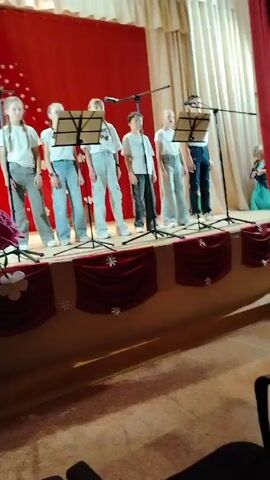 Seven children are standing in a line on a stage singing. They are positioned behind music stands and microphones. They all wear white shirts and jeans or gray pants. The stage is decorated with red fabric with white snowflake and flower details. A large bouquet of red roses sits to the side of the stage. The backdrop is a red curtain.