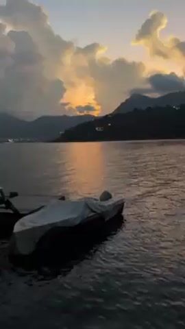 The video shows a picturesque lake scene at dusk. A boat covered with a light-colored tarp is docked at a small pier, floating in the dark water. In the distance, silhouetted mountains rise against a sky illuminated by the setting sun, which is peeking through the clouds and reflecting in the water. The clouds are thick and tinged with hues of orange and yellow. The scene is still and serene.
