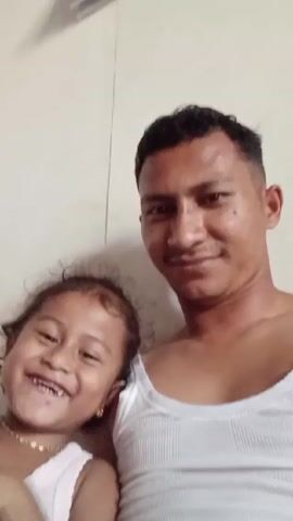 A man and a young girl are posing for a selfie, smiling and laughing. The man, possibly the girl’s father, is wearing a white ribbed tank top. He has short, dark hair and a light complexion. The little girl is wearing a similar white tank top and a gold necklace. She has dark hair and is holding her hand near her face, sometimes covering her mouth. The background is a plain white wall, with a glimpse of what appears to be a ceiling fan visible in the top left corner of the frame. The lighting suggests they are indoors. The interaction between the man and the girl is warm and playful.