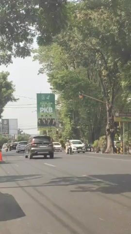 The video shows a street scene with cars and motorcycles. A large white truck is moving from right to left across the frame, obscuring some of the view. A billboard with green and white text and images of people is visible on the left side of the road. Trees line the street, and the sky is visible above. Traffic signals are present, with one appearing to be yellow. The perspective suggests the video is taken from a moving vehicle.