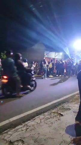 The video shows a nighttime street scene with a crowd of people. Lights, likely from a stage or event, are shining down, creating beams through a hazy atmosphere. Several individuals are walking and standing on the road and sidewalk. Some appear to be interacting with each other. Motorcycles are parked and one is in motion, with a rider visible. Buildings are present in the background. The overall atmosphere appears to be that of an outdoor gathering or event.