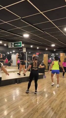 Several women are participating in a dance fitness class. They are dancing in a studio with a mirrored wall, a wooden floor, and a black ceiling with a grid pattern. One woman stands on a slightly raised platform and leads the class, facing the participants. The other women follow her movements. Most of the women wear athletic clothing. There is a "Salida" sign (Spanish for Exit) on the wall above the mirror. There are speakers and water bottles around the room.