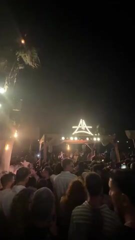 This video captures a nighttime outdoor event with a crowd of people gathered in front of a stage. The stage has a wooden structure and is illuminated by lights. A prominent logo, possibly representing a brand or artist, is displayed above the stage. Several palm trees are visible in the background, suggesting a tropical or warm-weather location. The atmosphere appears festive, with people dancing and enjoying the event. The lighting is dim, with focused illumination on the stage area.