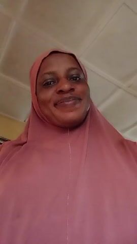 A person wearing a pink hijab is looking directly at the camera. The person is slightly smiling. The background consists of an off-white ceiling with white square panels. A small part of a yellow wall and a framed picture are visible in the background. The person is still throughout the video.