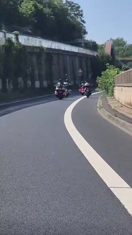 A group of three motorcycles are riding on a paved road. The road curves to the right, with a white line dividing the lanes. On the left side of the road is a steep embankment covered in greenery and a concrete wall. On the right side of the road, there are trees and a fence. In the background, an arched structure can be seen over the road. The sky is clear and blue, indicating it is daytime. The motorcycles are in motion, with the riders presumably operating them.