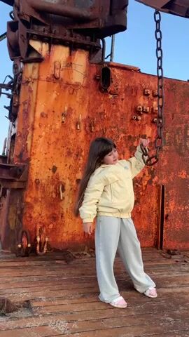 The video begins with a young girl with long, dark hair, wearing a light yellow jacket and light blue pants, standing on the deck of a rusty, dilapidated ship. She is holding onto a thick metal chain that hangs from above. The ship's surface is heavily corroded and orange, with wooden planks forming the deck. The scene then transitions to a wider view of the rusty ship, showing its various structures and a ladder. Following this, the video shifts to a beach scene at sunset. A sandy shore is visible in the foreground, with gentle waves lapping at the beach. In the distance, a body of water stretches towards a horizon with rolling hills or mountains. The sky is a gradient of blue, with touches of orange and pink from the setting sun, which is partially obscured by clouds over a small island or landmass in the distance.