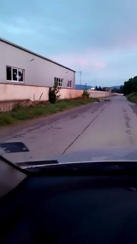 The video, captured from the dashboard of a vehicle, shows a road with a light grey building on the left. The building has a white base and several windows. A fence runs along the property line. The road surface is asphalt and appears somewhat worn, with cracks visible. There is grass and some vegetation along the side of the road. In the distance, the road curves to the right and slopes upwards. The sky is a pale blue with hints of pink, suggesting it is either dawn or dusk. There are a few dark objects on the road in the distance, possibly animals or debris. The camera is pointed forward, capturing the road ahead and the surrounding environment.