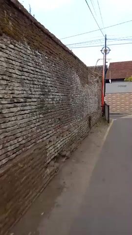 The video is shot from the perspective of someone riding a motorcycle, as indicated by the presence of a rearview mirror in the bottom left corner of the frame. The rider is traveling along a paved road, marked with a yellow line on the right edge. To the left of the road is a tall, old brick wall, showing signs of age and some mossy growth. The wall appears to be a boundary for a property. Further down the road, a metal railing, painted with alternating black and white vertical bars, is visible. Behind the railing, a property with a tiled roof and a modern building is partially visible. A red and white flag is attached to a pole near the property entrance, and an overhead utility line stretches across the top of the frame. The overall scene depicts a street in what appears to be a residential or mixed-use area, possibly in a warm climate given the vegetation.