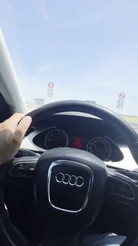 The video shows a first-person view from inside an Audi car driving on a highway on a sunny day. The driver's hand is visible on the steering wheel, which features the Audi logo. The dashboard shows the time as 13:42, temperature at 19 degrees, and speed around 75 km/h. A large truck is driving ahead on the highway, and the surrounding environment appears to be flat land. The sky is clear and blue with a few scattered clouds. The video seems to have been taken while the car was in motion.