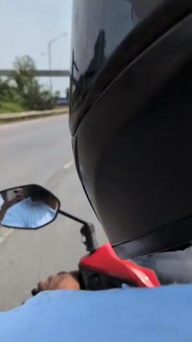 The video begins with a close-up shot of the back of a person's head wearing a helmet, with a rearview mirror of a motorcycle in the foreground. The mirror reflects a person wearing a light blue shirt, holding a smartphone, and presumably operating the motorcycle. The motorcycle is traveling on a paved road, with traffic visible in the distance. The road is bordered by a guardrail and vegetation on the left, and a construction site with concrete structures and shipping containers on the right. In the background, an elevated structure, possibly a bridge or highway under construction, is visible, along with utility poles and trees. The sky is partly cloudy. The video continues to show the motorcycle moving forward along the road, with the elevated structure and surrounding landscape passing by. The perspective shifts slightly as the motorcycle navigates the road, and the construction site and distant vehicles are intermittently visible.