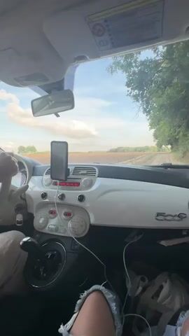 The video shows the interior of a white Fiat 500 car driving on a rural road. The dashboard is visible, with a smartphone mounted on the windshield. A person's hands are on the steering wheel, and another person's legs are visible in the passenger seat. The exterior view through the windshield shows a landscape with fields and trees. The lighting suggests it is daytime, possibly late afternoon.