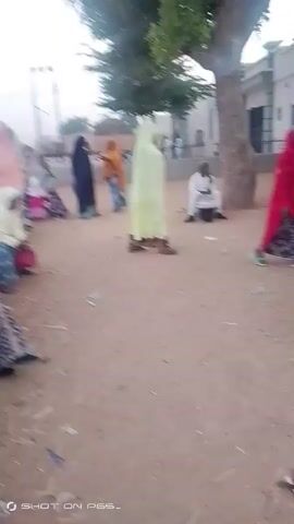 The video depicts an outdoor scene with several people, predominantly women wearing traditional clothing and head coverings, gathered in a dusty area. Some are sitting while others are standing and moving around. A man is seen riding a motorcycle in the background. There are buildings, trees, and a utility pole visible in the background, indicating an inhabited area. The overall atmosphere suggests a public gathering or a community space. The video quality is somewhat grainy. The text "SHOT ON P65" is visible in the lower left corner of the frame.