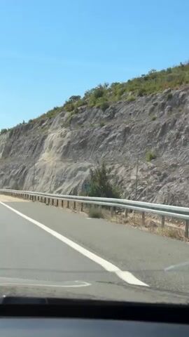 The video shows a view from inside a car as it travels on a highway. On the right side of the road, there is a steep rocky hillside covered in sparse vegetation. A metal guardrail runs along the edge of the road. Traffic signs are visible on the side of the road, including one with the number 228. In the distance, other cars are driving on the highway, and tall lampposts line the median. The sky is clear and blue.