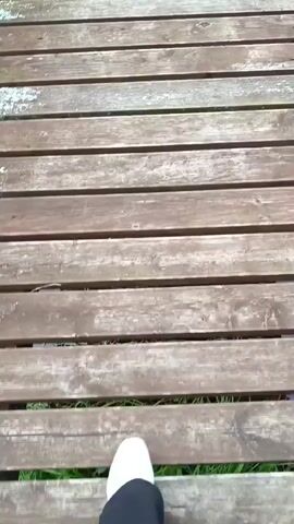 A first-person perspective video shows feet walking on a wooden deck. The deck appears to be made of weathered wooden planks, some of which are slightly grayed and showing signs of wear. Gaps between the planks reveal green grass below. The person is wearing white sneakers and dark pants, and they are walking forward across the deck. The camera angle is looking down at the person's feet and the deck surface.