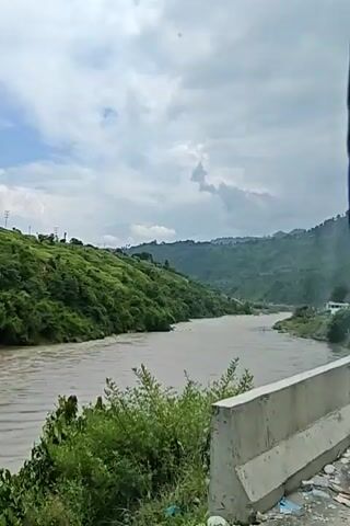 This is a video of a wide, muddy river flowing through a valley. The riverbanks are lined with lush green trees and vegetation. The sky is cloudy with patches of blue. In the foreground, there is a concrete barrier. The camera is moving, panning slowly to the right as it captures the scene. The video is likely taken from a moving vehicle, possibly a car or bus, given the perspective and slight vibration. The overall impression is one of a scenic, natural landscape.