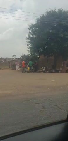 The video captures a drive through a rural or semi-urban street in Hoss, Nigeria, showing various everyday scenes. The time of day appears to be daytime, under an overcast sky.

The video begins with a shot of a man standing prominently in the middle of a dirt road, wearing a patterned shirt and looking to his left, possibly addressing someone or observing something. A large tree is in the background, and several simple buildings with corrugated iron roofs line the street.

As the vehicle moves, a woman in a yellow shirt and red skirt walks along the side of the road. The camera then pans across a row of small, open-front shops or stalls. Inside or in front of one of these shops, people are visible sitting. Two signs on the shop are readable: one clearly says "CLIPS," and another "CLIPS FRESH." A partial sign on an adjacent shop might read "POS."

Further along, a more dilapidated building with a blue door is briefly shown. The video then transitions to another small shop with a corrugated iron roof, where several colorful buckets are displayed outside. Two men are seen standing near this shop, and a motorcycle is parked in front of it.

Throughout the video, the environment is characterized by dirt roads, simple structures, and people going about their daily activities, reflecting a typical scene in a Nigerian community.