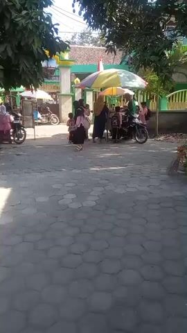 A group of children in pink and black uniforms are standing near a colorful umbrella. A woman in a hijab is walking towards the camera. There are motorbikes parked in the background, and a building with a green and white fence. The ground is paved with hexagonal tiles. The scene appears to be outside a school during the day.