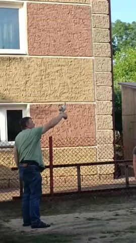A man in a green t-shirt and blue jeans stands in front of a building with textured walls. He is holding a pigeon in his outstretched right hand. The man appears to be looking up at the pigeon. There is a fence and some trees in the background.