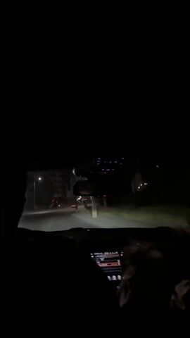 The video shows the view from inside a car at night. The dashboard and a rearview mirror are visible.  There are at least two people in the car, their silhouettes are visible in the foreground.  The car is driving down a street with streetlights and buildings visible on either side. Traffic lights are also visible in the distance. One person in the passenger seat has their hand raised and is making a gesture.