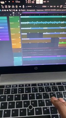 A close-up shot shows a person's hand interacting with the keyboard of an HP laptop. The laptop screen displays music production software, likely FL Studio, showing a "Playlist - Arrangement" view. Multiple tracks are visible, labeled "Track 1" through "Track 11," containing various audio clips and patterns with names like "HAT," "SATHAT," "PPSSattern 4," "KK," and "Mac Miller 110 bpm (consolidated)." A time marker in the software reads "1:02:13." The person's index and middle fingers are positioned over the 'G' and 'H' keys of the black QWERTY keyboard, suggesting typing or music control. The HP logo is visible on the laptop's lower bezel.