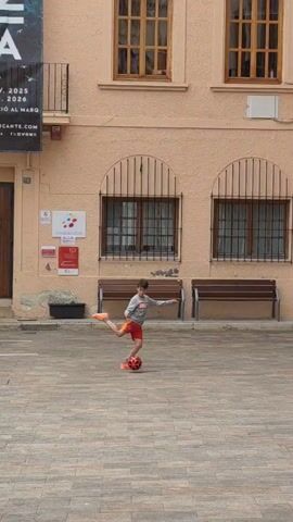 A young boy is playing with a red soccer ball in a brick-paved courtyard. He is wearing a gray sweatshirt, red shorts, and orange sneakers. The boy is kicking the ball and trying to keep it in the air. The courtyard is surrounded by light-colored buildings with windows and benches. A poster hangs on one of the buildings. The sky is overcast.