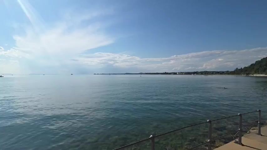 The video shows a panoramic view of a large body of water, likely a lake or the sea, under a bright, partly cloudy sky. The sun is shining, casting rays that create a lens flare effect on the left side of the screen. The water is a clear, turquoise color, and the bottom is visible in some areas, showing pebbles and rocks. Small ripples on the water suggest a gentle breeze. In the distance, there are several boats, including a small white boat and what appears to be a sailboat. Along the far shoreline, there is a line of trees and some buildings, suggesting a populated area. In the foreground, a black metal railing with a grey concrete walkway runs along the edge of the water, indicating a pier or promenade. The overall atmosphere is serene and summery.