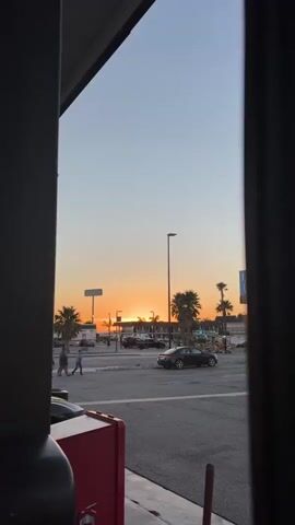 The video captures a sunset scene viewed through a frame of dark vertical structures on the left and right. The sky transitions from a pale blue at the top to a vibrant orange band near the horizon, where the sun is setting behind a low-rise building with palm trees in front of it. A street light stands tall in the middle ground. In the foreground, a street or parking lot is visible, with several cars parked and driving. A group of four people are walking across the street from left to right. A red object, possibly a dumpster or receptacle, is partially visible in the lower left corner. The lighting suggests it is late afternoon or early evening.