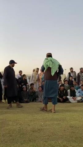 A group of people are gathered on a grassy field. In the center of the frame, a man in a blue tunic and brown vest is speaking to the crowd. He is wearing a light green scarf around his shoulders and a patterned cap. Another man in a dark tunic and cap stands facing him on the left side of the frame. The rest of the group consists of men, women, and children, many of whom are seated on the grass. The background shows a clear sky and distant structures. The lighting suggests it is either early morning or late afternoon.