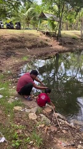 A man and a young child are by the edge of a pond. The man is wearing a maroon t-shirt and a black baseball cap, and is kneeling. The child is wearing a maroon t-shirt and is sitting. The man is pointing at the water, and the child is holding a stick and poking at the water. The pond is surrounded by dirt and some rocks. There is vegetation in the background, including trees and palm trees. There is also a small wooden shelter in the background. The overall scene appears to be outdoors in a natural setting.