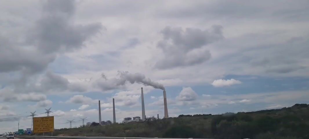 The video shows a view from inside a moving car, focusing on an industrial landscape. In the background, four tall chimneys stand prominently against a cloudy sky, with smoke billowing from one of them. Closer to the road, a yellow highway sign is visible, displaying directions in Hebrew, Arabic, and English, indicating an exit for "To OR AQIVA" at the "QESARIYYA INTERCHANGE." In the foreground, low-lying vegetation and a few electrical pylons are also visible.