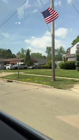 This media file captures a street-level view, likely from inside a vehicle, in Warrensville Heights, United States of America. The scene depicts a bright, sunny day with scattered clouds. The primary setting appears to be a commercial area with a few businesses and a residential feel to the surrounding trees and lawns.

The foreground shows a paved road with a curb, a storm drain, and a manhole cover. Beyond the road, on the left side of the frame, is a building that appears to be an auto repair shop or service center. A prominent sign for "AAA Approved Auto Repair" and "Center Hts Service" is visible. Several cars are parked in front of this building and in a lot adjacent to it, including SUVs and sedans.

To the right of the auto repair shop, the scene continues with more greenery and trees. A wooden utility pole stands prominently in the middle ground, with an American flag attached to it, fluttering in the breeze. Further down the street, a "STOP" sign with an arrow pointing right is visible. The overall atmosphere is that of a typical suburban commercial street on a pleasant day. No people are clearly discernible in the media.