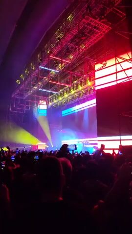 The video captures a concert scene from the perspective of a person in the audience. The view is looking towards a stage with a large rigging structure overhead, filled with various lighting fixtures that project beams of colorful light in yellow, blue, purple, and red. A large crowd of people is visible, with many holding up their phones to record the event. The environment suggests an indoor venue, possibly an arena or a large hall designed for concerts. The lighting creates a vibrant and energetic atmosphere, characteristic of a live music performance. The focus is on the stage and lighting, with the audience's faces mostly obscured.