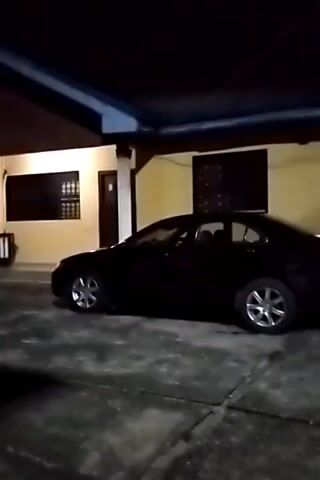 The video depicts a dark, nighttime view of a one-story building with a car parked in front. The building is light colored with dark windows and doors, supported by white columns. The car is a dark sedan with shiny hubcaps. The camera moves slightly, panning across the scene. The overall lighting is dim.