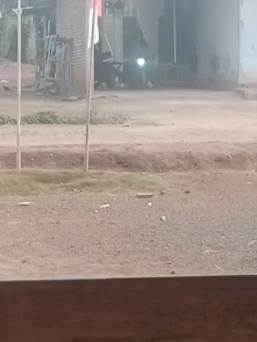 A person wearing a headscarf and dark clothing is riding a red and black motorcycle on a dirt road. The motorcycle has its headlights on. The person is riding past a white building with a green-trimmed window and some trees. There is also a flagpole with what appears to be an Indonesian flag. The ground is covered in dirt and gravel.